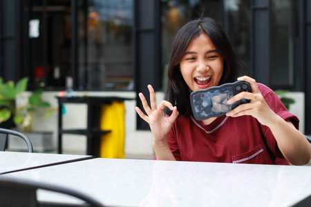 pretty asian woman playing game online using smartphone showing ok symbols sitting in outdoor cafeの写真素材