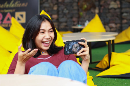 attractive asian woman excited playing online game using smartphone with raised hand sitting in...