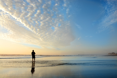 Man walking on beach at sunrise, beautiful cloudy sky reflected on the beach, Buildings, hotels in the background.  Jacksonville, Florida, USA.の写真素材