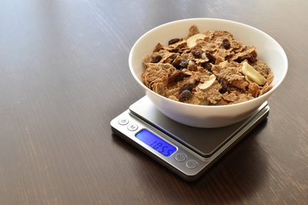 Fruit and fiber cereal in a white bowl on a kitchen scale on wood table with copy space on the left side.の写真素材