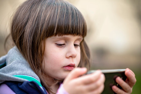 Cute little girl holding smartphone in one hand, watching videos outdoors, addiction conceptの写真素材