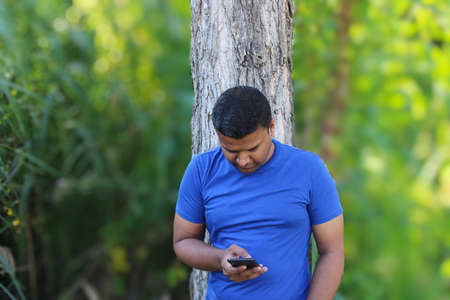 young man in blue t shirt standing with tree using cellphone outdoor in park