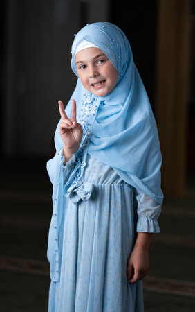 A happy Muslim child girl in hijab showing hand gesture v sign for victory or peace isolated portrait.の写真素材