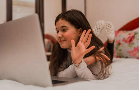 A happy teenage girl waving hand greeting gesture hello on video call while comfortable lying on the...
