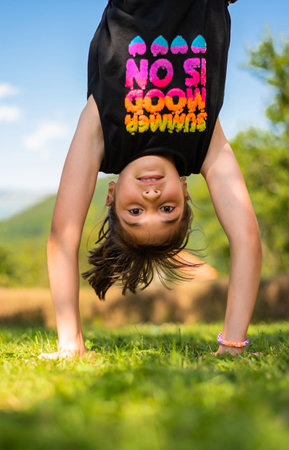 Happy kid girl having fun standing upside down on her hands on grass in the summer park.