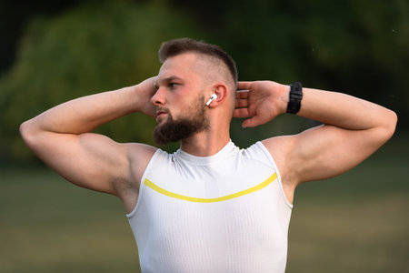 A young fit man doing stretching exercises before running or workout in a park, he listens to music...