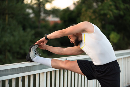 A young fit man is doing stretching exercises before running or workout on a city bridge, Outdoor fitness preparation, active lifestyle concept.の写真素材