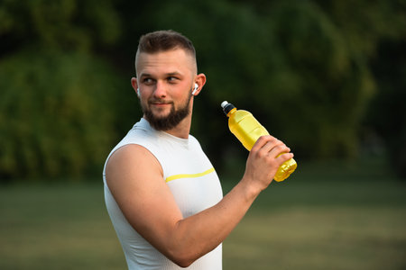 A young fit man listening to music on wireless headphones and drinking water or nutrition juice from a bottle after a run or workout in a park, active healthy lifestyle.の写真素材