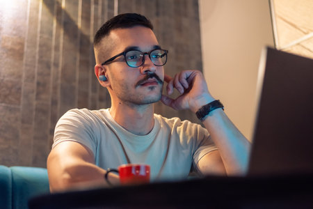 Focused young man wearing glasses and wireless earbuds, watching a webinar, having a video call with a client or a business conference meeting on a laptop in a modern interior with ambient light.の写真素材