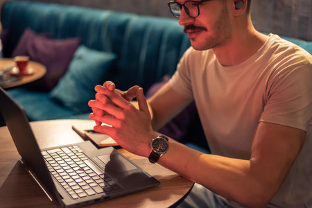 A happy young man working in a modern cafe, wearing glasses and wireless earbuds, having a video call with clients on a laptop, concept of remote work, freelancer lifestyle or online learning.の写真素材