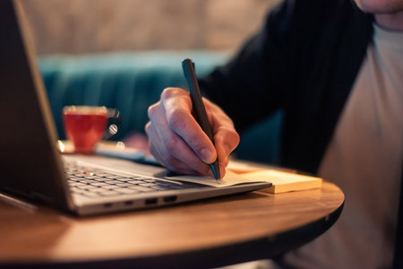 Close cropped image of a business man writing in a sticky note while working on a laptop or a male student studying in a modern interior. Concept of a business planning, strategy and online learning.の写真素材