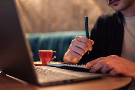 Close cropped image of a man writing in a notebook while working on a laptop or a male student studying in a modern interior. Concept of a business planning, strategy and online learning.の写真素材