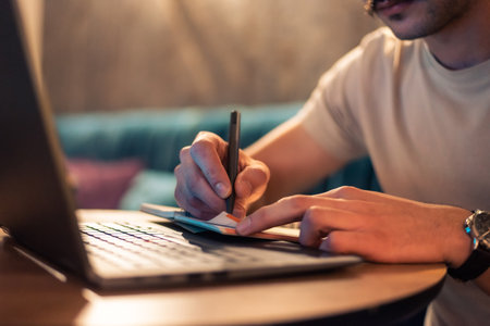 Close cropped image of a man writing in a notebook while working on a laptop or a male student studying in a modern interior. Concept of remote work, business planning, strategy and online learning.の写真素材
