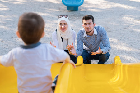 A happy Muslim family is playing together on the playground in the park, mom and dad are waiting with outstretched arms for the child as he descends from the slide.の写真素材