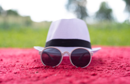 A fashionable white hat with a black band and round sunglasses placed on a red textured blanket....