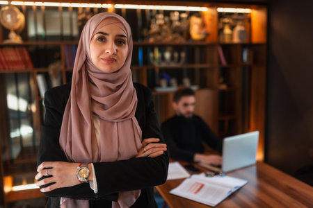 Confident businesswoman wearing a hijab standing with arms crossed in a modern office, a male colleague is working on a laptop in the background. Portrait of a business Muslim woman in the workplace.の写真素材