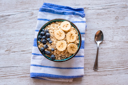 Top view of a porridge oatmeal with fresh fruit, a healthy breakfast bowl with oats, banana slices, and blueberries on a striped kitchen towel and rustic wooden table, with a spoon on the side.の写真素材