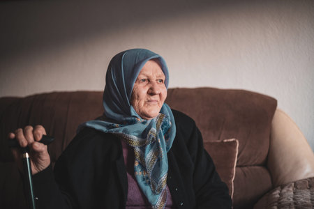 Portrait of an old Muslim woman with her walking stick at home wearing traditional hijab, looking away.の写真素材