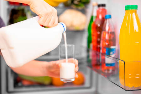 A girl pours milk from a bottle into a glass against the background of an open refrigerator with food. Close-up, small depth of field bright lightingの写真素材