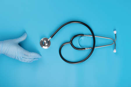 A doctor's hand in a medical glove reaches for a stethoscope on a blue background. The concept of diagnostics and treatment of diseases of the respiratory organs and heart.の写真素材
