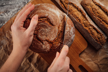 Women's hands cut fresh rye bread with a knife on a wooden table sprinkled with flour and covered...