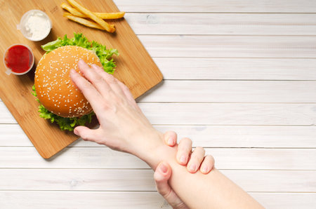 The girl shows willpower by holding with her left hand, her right hand that reaches for a hamburger. Diet and healthy nutrition, top view.の写真素材