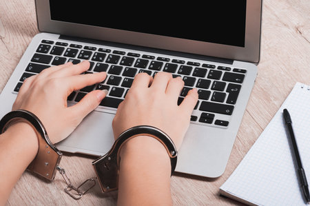 A girl in handcuffs is sitting at a laptop. The concept of censorship or restriction of freedom of speech or punishment for disclosure of confidential information. Very close-up.の写真素材