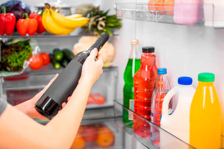 Girl holding a bottle of wine against the background of an open refrigerator with food, close-up, shallow depth of field bright lightingの写真素材