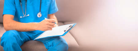 A doctor in a blue uniform with a stethoscope sitting on the couch records anamnesis or prescribes a course of treatment. Pastel colors are a place for your textの写真素材