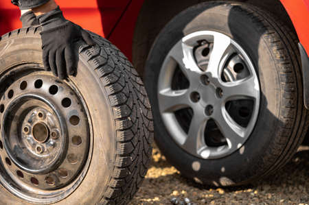 A man rolls a wheel with a studded tire on the background of a red car with a summer tire. The concept of replacing the summer wheel with a winter wheel with your own handsの写真素材