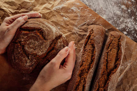 Women's hands put fresh rye bread on a wooden table sprinkled with flour and covered with parchment paper. The concept of making natural homemade bread or bakery. Rustic style brown tones.の写真素材