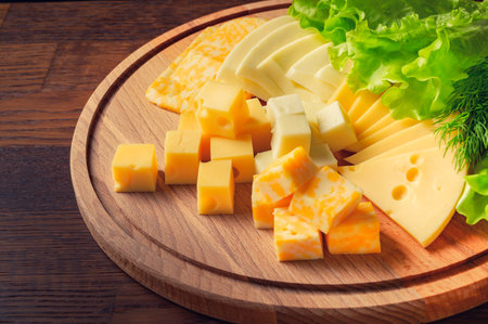 Square pieces of cheese with herbs on a round wooden board.の写真素材