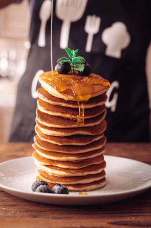 Stack of fresh homemade pancakes on a platter with berries and fresh honey. Cook in an apron in the background.の写真素材
