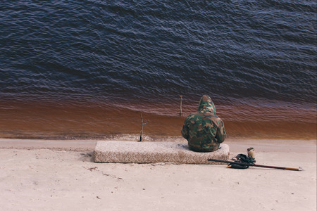 The sitting fisherman with a fishing tackle on lake. in camouflage suit.の写真素材
