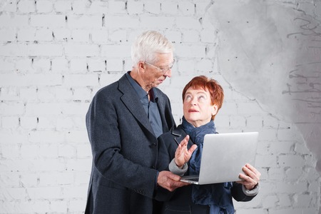 Old people hold a laptop and communicate through the Internet. Happy smiling grandpa grandma couple standing cuddling together isolated on white brick backgroundの写真素材