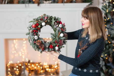 Young woman holding a Christmas wreath with fir branches for the holiday. The new year celebration. on the background of fireplace and Christmas tree, lights, garlands