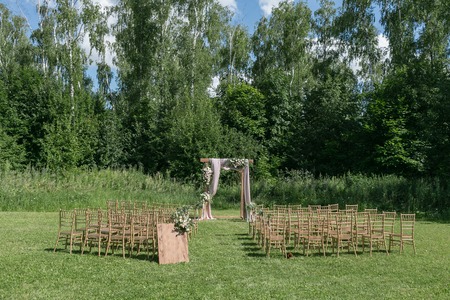 Beautiful setting for outdoors wedding ceremony waiting for bride and groom and guests. Wooden chairs decorated with flowers, are in the zone of the wedding ceremony in a forestの写真素材