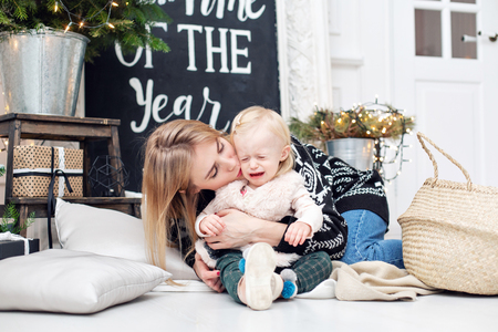 babys crying. Cheerful mom embraces her cute daughter . Parent and little child having fun near Christmas tree indoors. Loving family Merry Christmas and Happy New Yearの写真素材