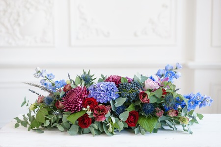 Elongated floral arrangement in vintage metal vase. table setting. Blue and red color. Gorgeous bouquet of different flowers. Bright room, stucco on the wallの写真素材
