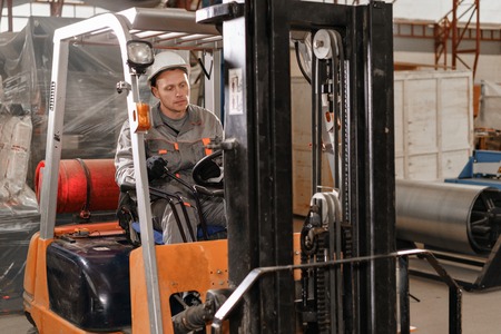 man driving a forklift through a warehouse in a factory. driver in uniform and protective helmet....