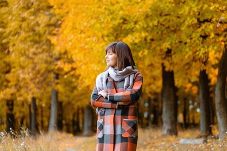 Portrait of young woman in fall forest. Brunette woman in autumn park with fashionable plaid coat and scarf.の写真素材