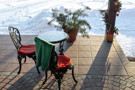 Cast iron chairs and tables with pillows and a blanket on restaurant terrace. Winter landscape of frosty trees, white snow and blue sky. Tranquil winter nature in sunlight in parkの写真素材