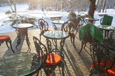 Cast iron chairs and tables with pillows and a blanket on restaurant terrace. Winter landscape of frosty trees, white snow and blue sky. Tranquil winter nature in sunlight in parkの写真素材