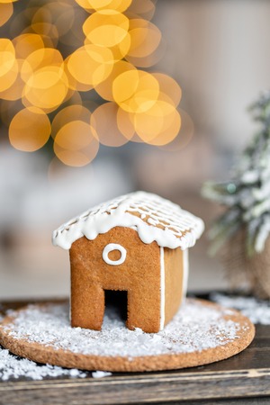 Gingerbread house on wooden table. Defocused garland lights on background. Christmas tree and Holiday mood. Morning in the bright living room.の写真素材
