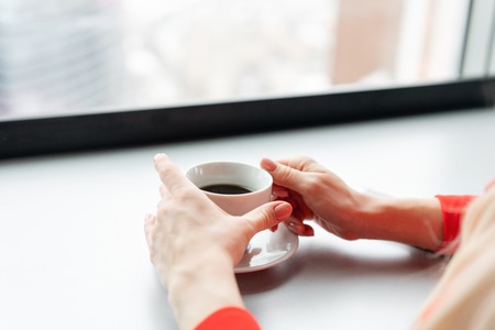 Cup of hot drink in hand close-up. Woman in red coral business suit drinks coffee at a high table near the window. Young blonde businesswoman in the office in high floor in front of the window.の写真素材