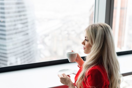 Woman in red coral business suit drinks coffee at a high table near the window. Young blonde businesswoman in the office in high floor in front of the window.の写真素材