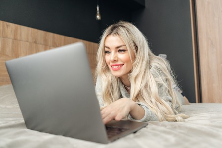 portrait of a girl behind a laptop screen. Beautiful blonde woman lying on the bed and looks at the laptop screen. Blogging, browsing internet, chatting. In a warm cozy sweaterの写真素材
