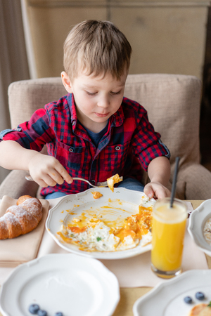 Funny and happy boy having Breakfast. Light brunch near window in a cafe. Croissants, omelet, orange juice and many different dishes on the table in the cafe.の写真素材