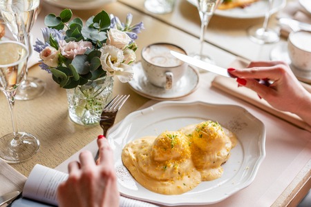 Close up Woman eats Egg Benedict on warm toast brioche. Two eggs delicious hollandaise sauce. Light morning Breakfast on table in restaurant.の写真素材