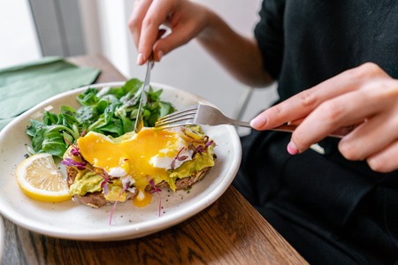 Young woman eat breakfast with knife and fork, Morning in cafe. Healthy breakfast with wholemeal bread toast with avocado, poached egg with green saladの写真素材